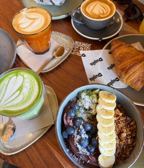 Breakfast in Bilbao, Spain: Açai bowl topped with blueberries, bananas, kiwi, cooconut, and granola, with a croissant and matcha latte on the side. Friends ordered cappuccinos and bagel sandwiches.